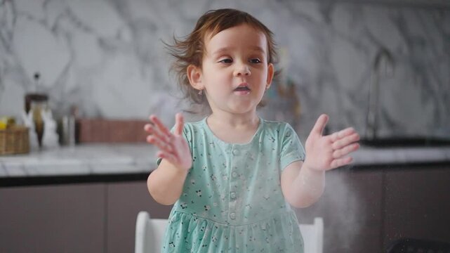 Little girl claps hands, flour flies around her. Her eyes show delight, not annoyance. Kitchen background is marble and clean. Perfect for playful child moments or cooking fun