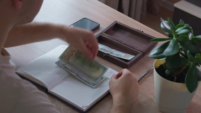 saver separates cash dollars inside wooden tray as part of structured reserve fund. responsible person arranges stacks of paper money near potted plant and smartphone for reliable emergency cash.