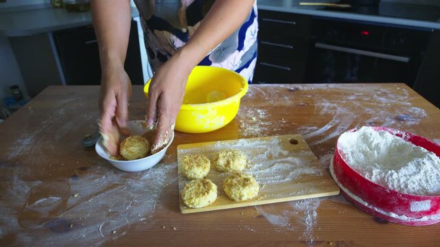 Hands shape dough on floured wooden board. Yellow bowl holds crumbs for coating. Flour bowl rests nearby, dusted surface. Joyful hands create golden treats