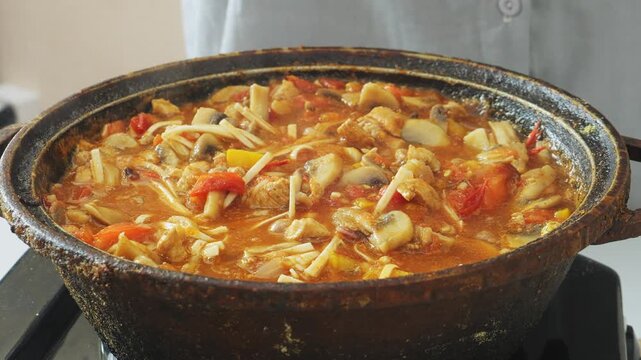 Vegetables simmer in a deep cast-iron skillet with chicken and udon, stirring with a spoon. Medium shot.
