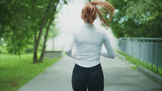 Woman runs through park path. Hair flows behind her as she moves. Trees line the walkway softly. Green grass borders the path. She wears fitted athletic gear. Ideal for fitness or wellness content