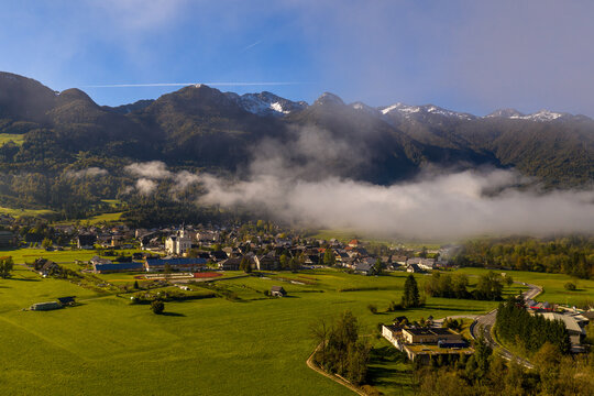 Aerial view of the town nestled in a green valley with low-hanging clouds, surrounded by the Julian Alps under a clear blue sky in Bohinjska Bistrica, Radovljica, Slovenia.