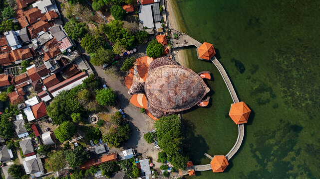 Aerial view of Kura-Kura Ocean Park with its turtle-shaped building, pedestrian walkway, and coastal trees along the clear green sea in Jepara, Central Java, Indonesia.