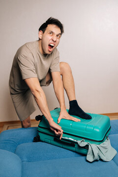 Funny excited angry traveler male struggling to zip overpacked green suitcase for a trip, frowning and making a funny face, experiencing frustration and packing difficulty for vacation travels.