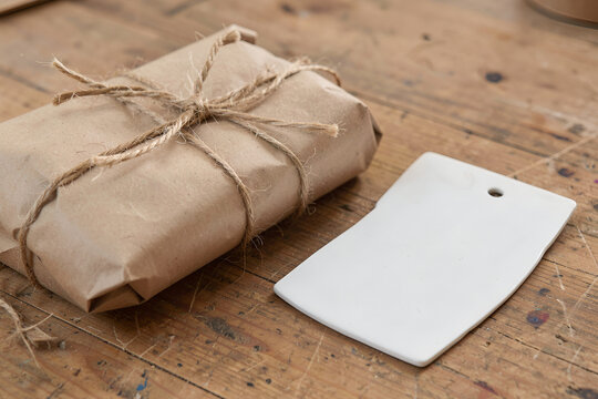 Handmade kraft-paper parcel tied with twine beside a blank ceramic tag on a wooden worktable