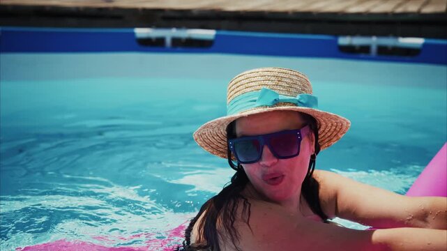 Woman smiles, wearing hat and sunglasses in pool. She rests on a pink float, enjoying sunny day. Water ripples gently around her as she relaxes. Perfect for vacation, pool party