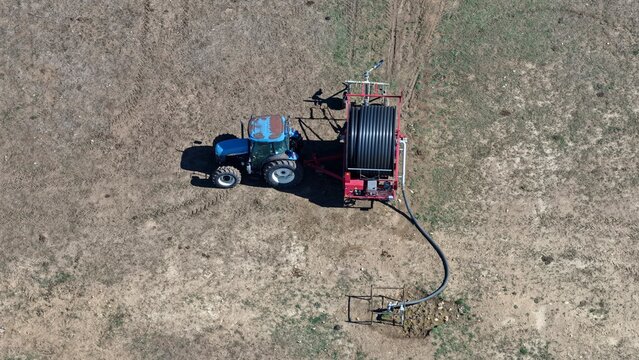 Aerial view of a blue tractor pulling a large irrigation hose reel across a dry, brown agricultural field in Niwot, Colorado, United States.