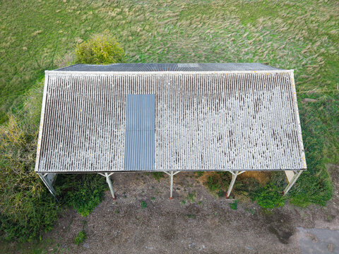 Drone view of an old, asbestos-roofed hay and stray shelter used to keep hay dry as seen on a dirty farm. A dairy pasture surrounds the building.