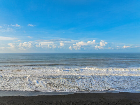 Aerial view of Pantai Pasir Besi with its distinctive black sand beach, crashing white waves, and a clear blue sky over the ocean in Cibitung, West Java, Indonesia.