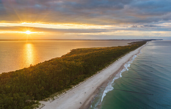 Aerial view of Hel Peninsula at sunset with a sandy beach, dense forest, and wooden groynes extending into the Baltic Sea in Chalupy, Pomeranian Voivodeship, Poland.