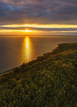 Aerial view of the Hel Peninsula at sunset, with golden light reflecting on the Baltic Sea, dense pine forests, and a dramatic sky in Chalupy, Pomeranian Voivodeship, Poland.