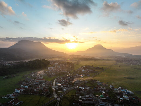 Aerial view of Kadungora village nestled among lush rice fields and mountains at sunset in Kadungora, West Java, Indonesia.