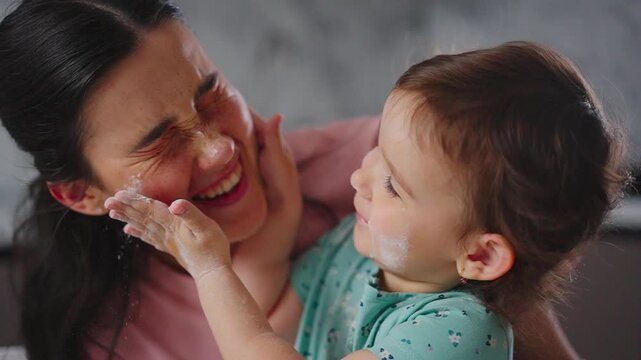 Mother laughs as child playfully smears face with cream. Child giggles, hands smeared with white substance. Tender moment shows joyful parent-child bonding. Perfect for family, parenting