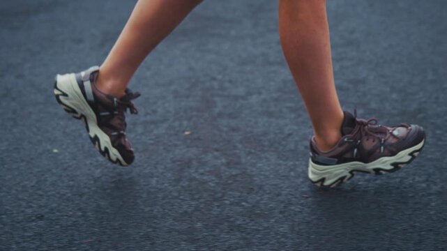 Active runner strides on asphalt road. Athletic legs move with purpose, steady rhythm. Dark, chunky shoes grip pavement with force. Energy flows through each determined step