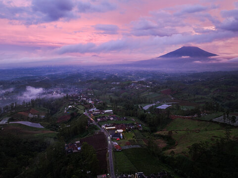 Aerial view of Darajat Pass at twilight with a purple sunset over the mountain, lush hills, agricultural fields, and a winding road in Kecamatan Pasirwangi, Jawa Barat, Indonesia.