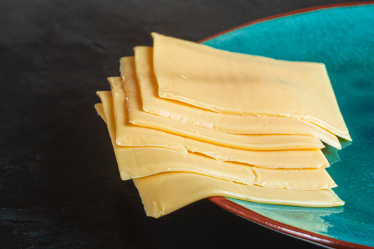Stack of processed cheddar cheese slices on turquoise plate over dark background. Dairy product, sandwich ingredient, burger cheese, fast food prep and culinary staging concept.