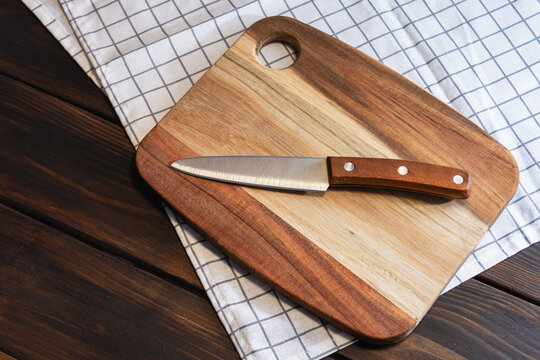Stainless steel utility paring knife lying on wooden cutting board with checkered napkin on dark wood table. Kitchen tools, culinary preparation, food staging and cooking accessories concept.
