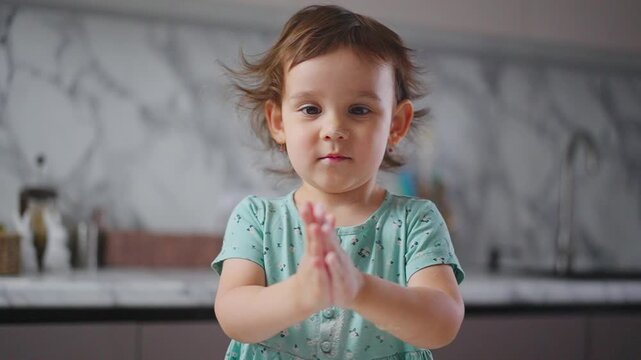 Child claps hands in kitchen. Child faces camera with curious expression. Child wears light green patterned shirt. Kitchen has marble backsplash. Ideal for family, children, playful moments