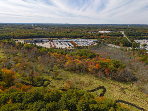 Aerial view of a commercial shopping center with a large parking lot surrounded by autumn forests and a winding stream in Rochester, Massachusetts, United States.