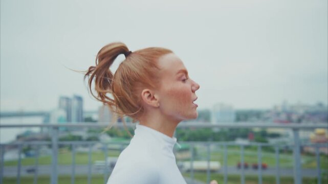 Red-haired woman runs on urban rooftop. Wind tugs her ponytail, blowing her hair. City skyline visible behind metal railing. She moves forward with focus, steady pace