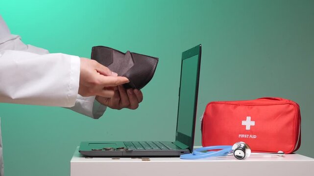 Person in white coat opening an empty wallet, demonstrating financial hardship and high medical costs with coins falling onto a laptop keyboard, a stethoscope, and a red first aid bag nearby