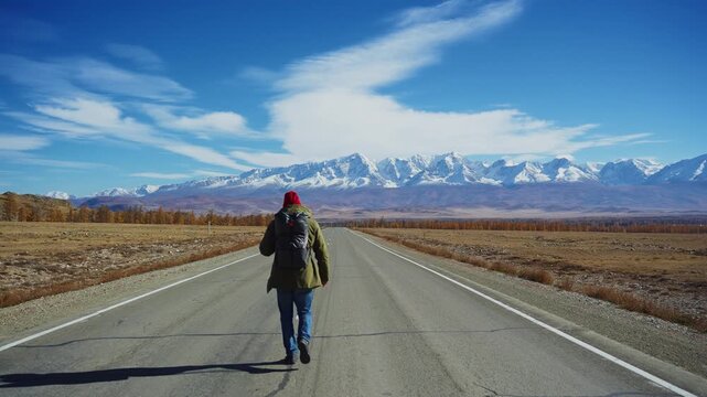 Lone figure walks down a vast, open road. Mountains stand tall, snow-capped and serene. Sky is bright blue with wispy clouds. Person wears a red hat and green jacket. Wind moves the figure's hair