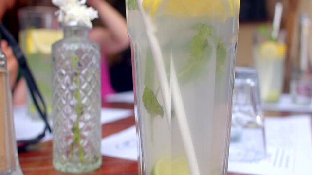 Tall clear glass filled with pale citrus drink, lemon and lime slices, mint leaves, and white straw on table in summer.