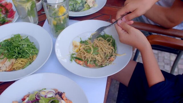 Overhead view hands twirling spaghetti with shrimp and leafy greens using fork and spoon on outdoor table during casual lunch.
