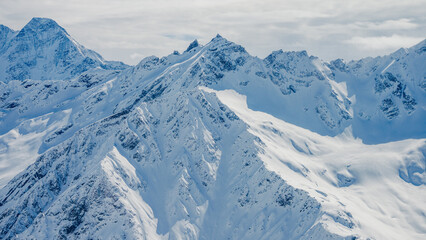 Aerial view of snow-covered Mount Elbrus and jagged Caucasus ridgelines under overcast sky © ossyffer