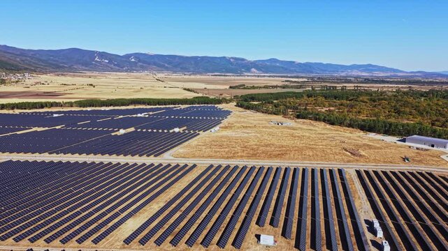 A large solar panel farm spreads across the ground in a wide, flat area. Green trees and distant mountains are visible under the clear blue sky.