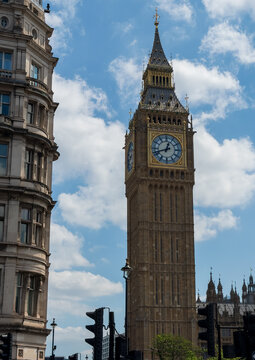 Big Ben Elizabeth Tower with gold and blue clock face beside a historic stone building and traffic lights under a cloudy blue sky in London