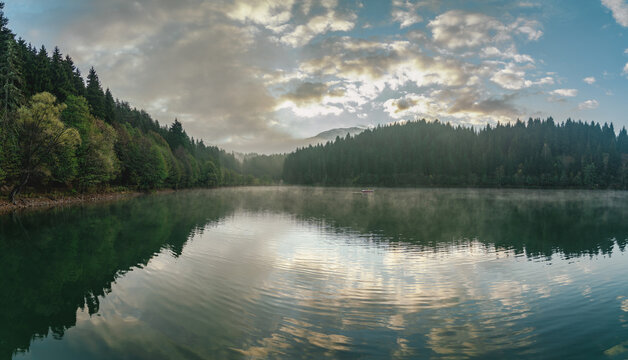 Karagol lake near Savsat, Artvin, Turkey, a panoramic view with clouds reflected on rippling water, mist on the surface and a small boat between the fir forests