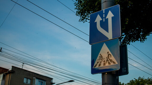 Traffic signs show directions for vehicles and cyclists at a busy intersection in a city. It is late afternoon, and the sky is clear
