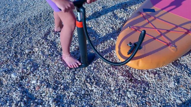 Woman inflating a stand up paddleboard on a pebble beach. Woman preparing for water sports, using a manual air pump to inflate her stand up paddleboard on a sunny pebble beach before paddling