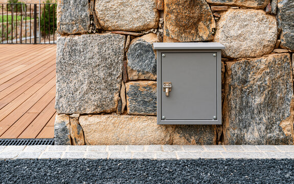 Grey electrical box mounted on textured stone wall. Outdoor utility junction box with metal latch on granite facade. 