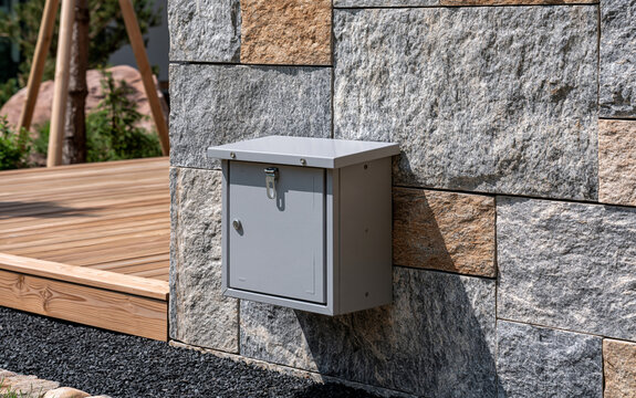 Grey electrical box mounted on textured stone wall. Outdoor utility junction box with metal latch on granite facade. 