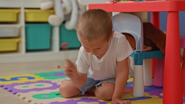 Boy covering mouth with hand while kneeling on foam number mat playing with wooden toys and sitting by red table and toy kitchen shelves in building; quiet curiosity.