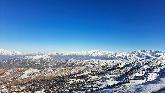 ​Wide-angle panoramic view of a majestic mountain range with snow-covered peaks under a clear vibrant blue sky. Breathtaking high-altitude winter landscape showcasing layered snowy slopes and mountain