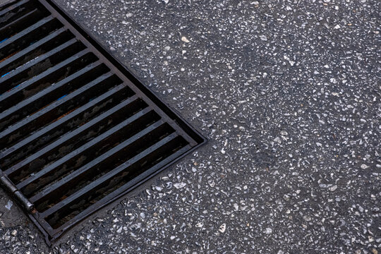 Storm drain grate on asphalt. A cast iron grate with parallel bars sits flush against a gritty aggregate road surface, viewed from above. Useful for infrastructure, drainage, or urban engineering.