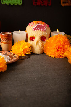Day of the Dead altar offerings with a sugar skull, clay plate with sea salt, traditional tequila shot, burning candles, and orange marigold flowers on a dark rustic table.