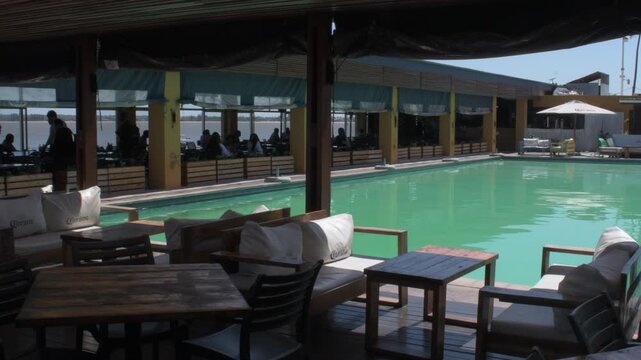 view of an open air seaside settlement and poolside lounge at the maputo waterfront featuring outdoor wooden seating, dining tables, and a turquoise pool overlooking the bay under bright daylight