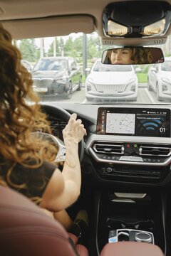 A woman with curly hair is focused on driving a car, checking the navigation screen. She is inside a modern vehicle parked among several other cars on a clear day