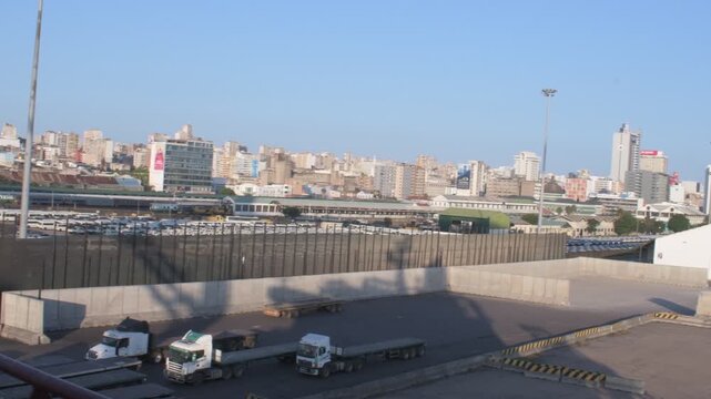 elevated panoramic perspective across the port of maputo logistics area showing heavy cargo transport trucks, storage yards, railway lines, and warehouse facilities fronting the city skyline under bri