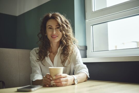 A woman with curly hair sits at a wooden table in a cafe, holding a cup of coffee. She smiles while looking out of a large window that lets in bright afternoon light