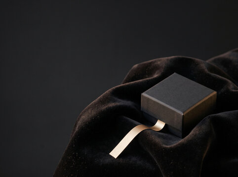 BlackFriday, Closed dark square gift container resting atop soft fabric drapes featuring an extended gold ribbon strand stretching across the textured surface with ample negative spac Luxury