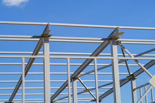 Steel frame structure of prefabricated building with square tubes, H-beams, horizontal beams. White metal framework close-up with bolted joints, support plates, column and roof beam connections.