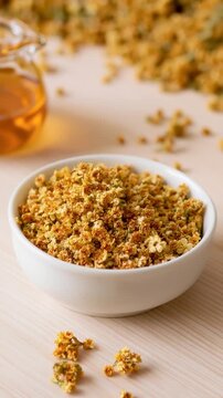 Dried golden osmanthus flowers in a white bowl beside a glass jug of amber syrup on light wood, evoking floral tea, sweetness and delicate scent