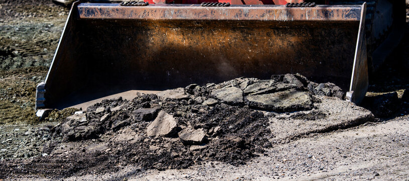 Asphalt removal on damaged road. Broken asphalt fills loader bucket. Asphalt chunks show rough texture. Excavator clears asphalt debris outdoors.