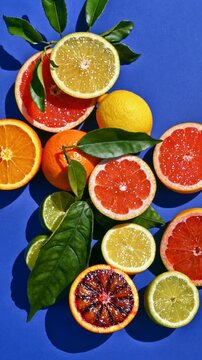 Colorful halved grapefruits, oranges, lemons and limes with glossy leaves arranged on a bold blue surface, top view of juicy fresh citrus