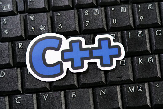 C++ programming language logo sticker placed on black laptop keyboard, blue plus signs on white cut out, closeup of computer keys with numbers and letters, software development concept